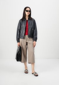 Giacca bomber in pelle nera, top rosso, pantaloni cropped beige, borsa nera e sandali decorati. La modella indossa occhiali da sole scuri. Sfondo semplice.