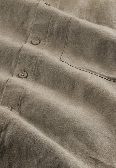 Beige linen shirt with a wrinkled texture, featuring buttons and a chest pocket. Fabric shows natural fibers and subtle variations in color.