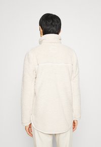 Pehmeä kermainen fleece-pullover takki korkealla kauluksella, jossa takana sauma- ja pyöristetty helmareuna. Pehmeä materiaali ja rento istuvuus.