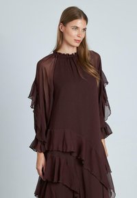 Langes, fließendes Kleid in dunklem Burgunderrot, mit gerafftem Stoff und durchsichtigen Puffärmeln. Der Ausschnitt ist gerafft für zusätzliche Details.