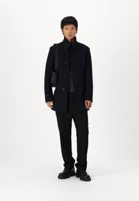 Navy wool coat with a high collar, button closures, and side pockets, paired with black trousers and shoes, worn with a crossbody bag.