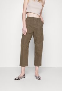 Kaki Cropped-Hosen mit tailliertem Schnitt, ausgestattet mit seitlichen Taschen und einer glatten Baumwolltextur. Kombiniert mit gemusterten flachen Schuhen.