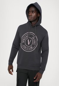 Tmavošedá mikina s kapucňou a veľkým okrúhlym logom v svetloružovej farbe. Má šnúrky a elastické manžety, vyrobená z jemného materiálu.