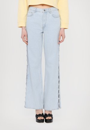 Lichtblauwe wijde jeans met zijknopen; denim materiaal; uitgerafelde zoom aan de onderkant; gestyled met zwarte plateausandalen.