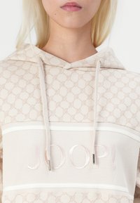 Beige Kapuzenpullover mit strukturiertem Muster, Frontlogo "JOOP!" in hellrosa Stickerei, mit Kordeln und horizontalen weißen Streifen.