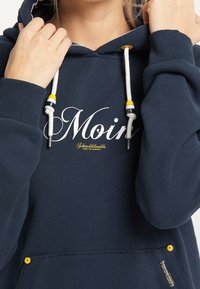 Navy hoodie with "Moin" text in white and yellow. Features adjustable flat drawstrings with colored accents and kangaroo pocket.