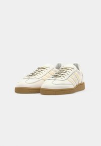 Scarpe da ginnastica bianche con tomaia in pelle, accenti beige, design a tre strisce, punta rotonda e suola in gomma. I lacci sono di un grigio chiaro.