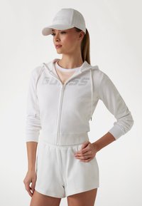 Bijela zip-up majica s kapuljačom s ukrašenim logom, u kombinaciji s bijelim kratkim hlačama. Uključuje bijelu kapu s istim logom. Glatka tekstura.