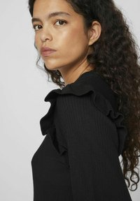 Melnā ribota džemperis ar viļņainiem akcentiem pie pleciem, bagātīgas tekstūras audums, pieguļoša silueta un smalki šuvumi.