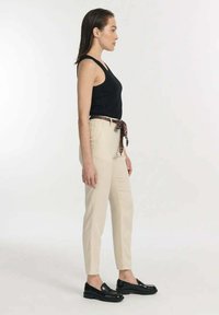 Beige schmal zulaufende Hose mit einem schwarz-braun gemusterten Gürtel, kombiniert mit einem schwarzen Tanktop und schwarzen Slippern. Glattes Material mit figurbetonter Passform.