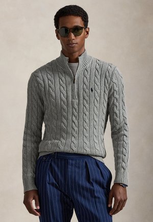 Homme portant des lunettes de soleil noires, un pull gris en maille torsadée à demi-fermeture éclair et un pantalon à fines rayures bleu, debout devant un fond neutre.