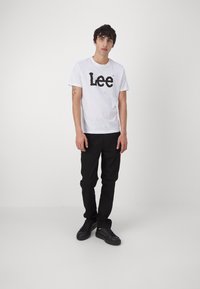 Hvit kortermet t-skjorte med svart "Lee" logo, kombinert med svarte bukser og svarte joggesko, stående mot en lys grå bakgrunn.