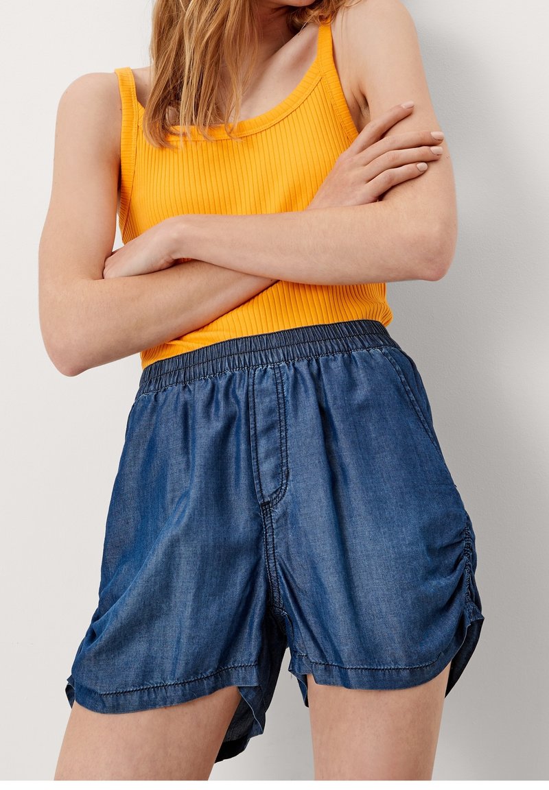 Shorts in denim con vita elastica, con arricciatura laterale, indossati con un canottiera gialla a coste. I pantaloni shorts sono di una tonalità blu medio.