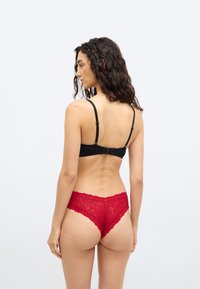 Γυναίκα με μακριά σγουρά μαλλιά, φαίνεται από πίσω, φοράει μαύρο σουτιέν και κόκκινο δαντελωτό εσώρουχο πάνω σε απλό ανοιχτό φόντο.