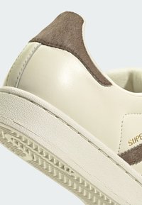 Krémovo kožená teniska s textúrovaným hnedým semišovým záhlavkom, gumovou podrážkou a viditeľným prešívaním. Názov značky je vyrazený zlatým písmom.