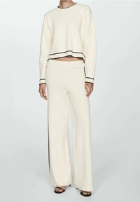 Donna che indossa un completo a maglia crema in due pezzi con rifiniture nere, composto da un maglione cropped a maniche lunghe e pantaloni a gamba larga, abbinato a tacchi neri open-toe.