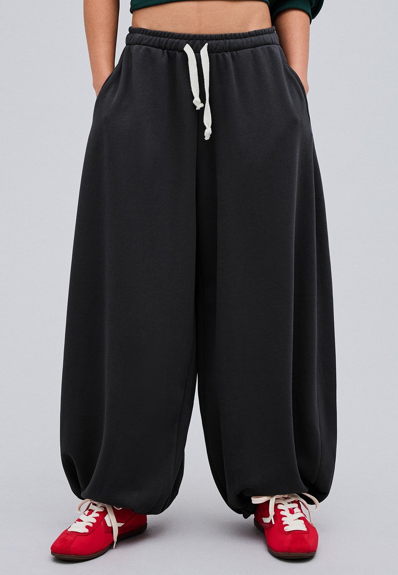 Personne portant un pantalon noir ample avec des cordons blancs et des baskets rouge vif, debout devant un fond uni.