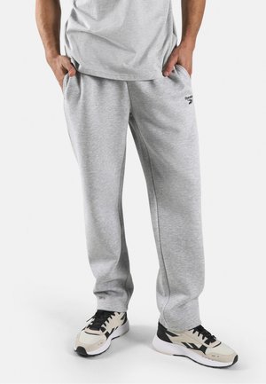 Man wearing light grey Reebok sweatpants and white, black, and beige sneakers, standing with hands in pockets against a white background.