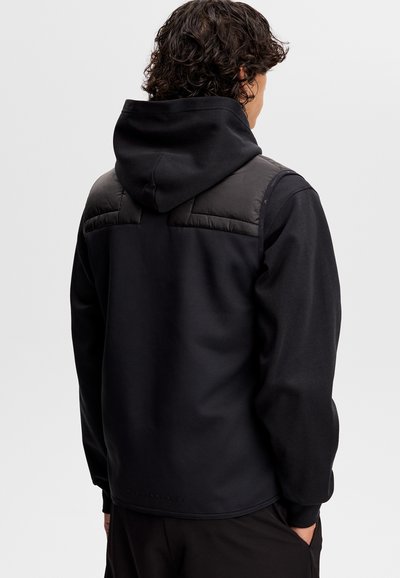 Mężczyzna z kręconymi włosami, ubrany w czarną bluzę z kapturem i puchową kamizelkę, stoi tyłem na jednolitym, jasnoszarym tle.