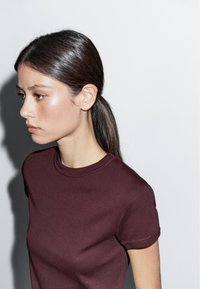 Bordeaux kortemouwen t-shirt van zacht katoen, met een ronde hals en een gladde textuur. Model met lang, steil haar in profiel.