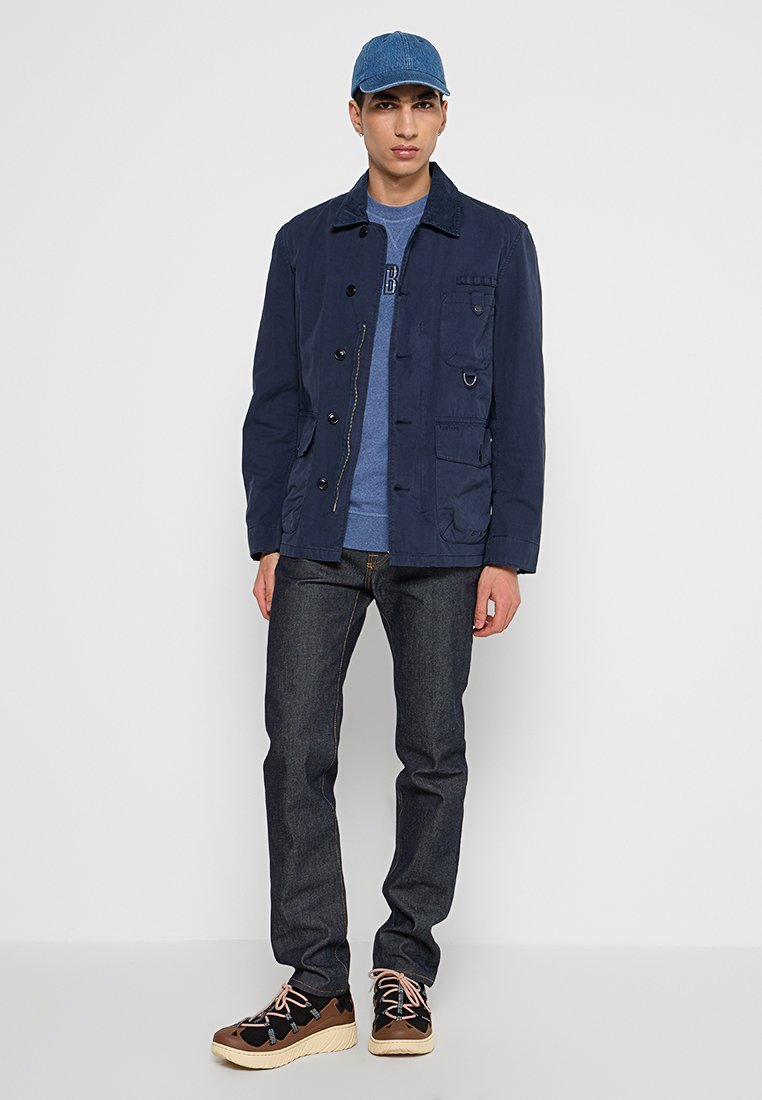 Giacca blu navy con chiusura a bottoni e tasche frontali, abbinata a una t-shirt blu, jeans scuri e sneakers marroni con lacci.