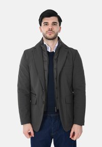 Giacca doppiopetto grigio scuro con taglio moderno, dotata di uno strato interno trapuntato. Tessuto strutturato e revers definiti. Bottoni e tasche visibili.