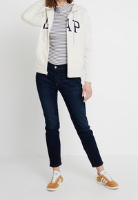 Sudadera de cremallera gris claro con el logo "GAP", cuello de tortuga a rayas, jeans de mezclilla oscura y zapatillas marrones y blancas. Tejido suave, ajuste casual.