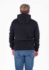 Persona con capelli biondi corti che indossa una felpa nera con cappuccio e jeans blu, in piedi di spalle su uno sfondo bianco.