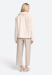 Veste en fausse fourrure crème avec un large col en shearling, coupe décontractée et une fermeture à un bouton à l'arrière ; associée à un pantalon beige clair.