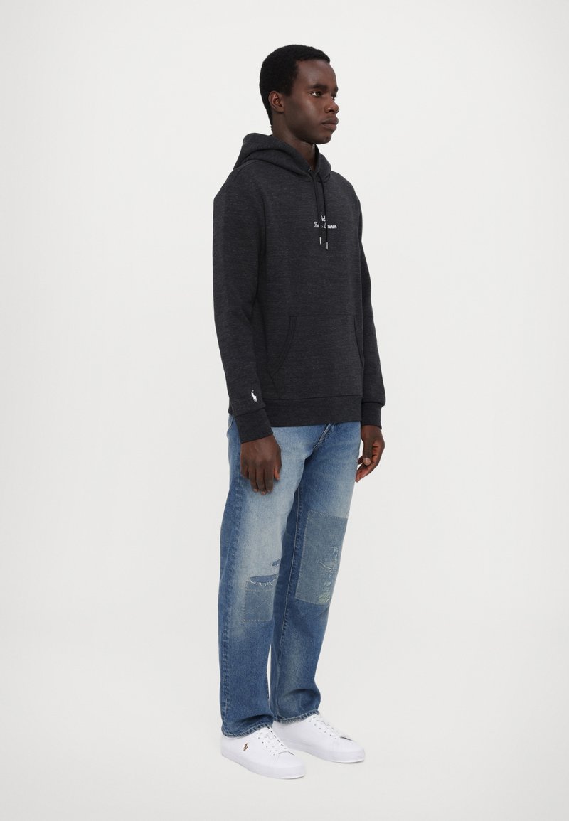 Schwarzer Kapuzenpullover mit Fronttasche und weißem Logo; hellblaue, ausgewaschene Jeans mit Patchwork; weiße Sneaker. Modell steht seitlich.