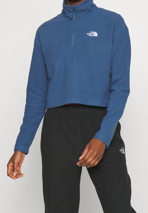 Person wearing a blue cropped quarter-zip fleece with white logo and black sweatpants featuring a white logo on the left thigh.
