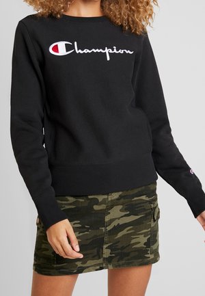 Vrouw draagt zwarte Champion sweater met wit en rood logo, gecombineerd met een groene camo cargo minirok tegen een lichtgrijze achtergrond.