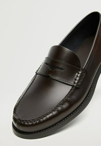 Tummanruskea nahkaloaferi, jossa on ommeltuja yksityiskohtia ja koristeellinen raita ylhäällä, esitettynä yksinkertaista valkoista taustaa vasten.