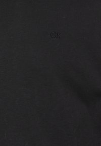 Schwarzes Baumwoll-T-Shirt mit kurzen Ärmeln, das ein erhabenes "CK"-Logo auf der Vorderseite zeigt. Glatte Textur und lässiges Design.