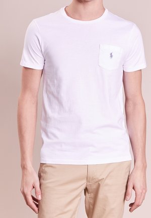 Man draagt een eenvoudig wit T-shirt met korte mouwen, een klein zakje en logo op de linkerborst, gecombineerd met beige broek.