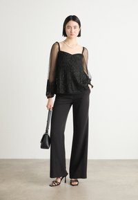 Schwarzes Paillettenoberteil mit durchsichtigen Ärmeln, kombiniert mit hochtaillierten schwarzen Hosen und schwarzen Riemchenheels; Modell hält eine kleine schwarze Tasche.