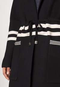 Schwarze Strickjacke mit weißen Streifen an der Taille und Taschen, mit einer Kordelzug-Verschluss und metallischen Akzenten an den Taschen.