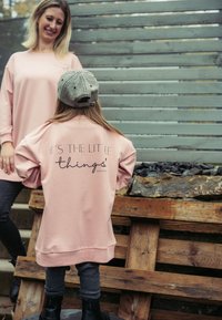Rosa übergroßer Sweatshirt mit schwarzen Text auf dem Rücken, kombiniert mit grauen Leggings und schwarzen Stiefeln. Der Hintergrund zeigt Holzpalette und einen Zaun.