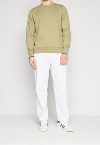 Felpa verde oliva con collo rotondo e polsini a costine, caratterizzata da un piccolo logo ricamato. Abbinata a pantaloni bianchi morbidi e sneakers.