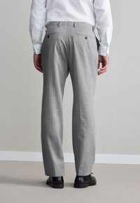 Pantaloni da uomo sartoriali grigi con tessuto strutturato, davanti piatto e doppie tasche posteriori, abbinati a eleganti scarpe nere lucide.