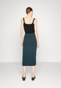Schwarzes Tanktop mit dünnen Trägern, kombiniert mit einem gerippten, türkisfarbenen Midirock aus strukturiertem Material und schwarzen Sandalen mit Blockabsatz.