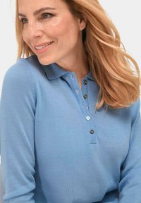 Lichtblauwe langmouw polo met een ribbelstructuur, voorzien van een knoopsluiting met twee donkere knopen en een enkele lichte knop.