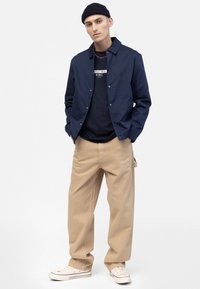 Chaqueta ligera de color azul marino con cuello y botones a presión sobre una camiseta oscura, combinada con pantalones de pierna ancha beige y zapatillas blancas.