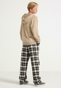 Lys blond person som står med hendene i lommene, iført beige hettegenser, svarte og hvite rutete bukser, og grå Converse-sneakers, på hvit bakgrunn.