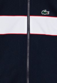 Veste zippée bleu marine présentant une bande horizontale blanche avec un contour rouge, et un logo de crocodile vert en haut à gauche.
