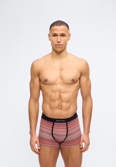 Homme musclé aux cheveux courts, debout face à l'avant, portant un boxer rayé multicolore avec une ceinture noire et le logo "Paul Smith".