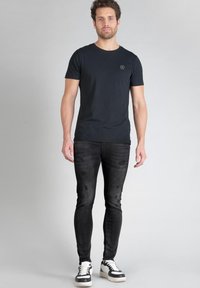 T-shirt noir à col rond avec un petit logo sur la poitrine, associé à un jean noir ajusté avec des détails déchirés et des baskets noir et blanc.