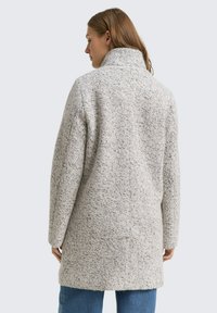 Manteau en laine texturée gris avec un col haut, coupe légèrement oversized, coutures visibles et design mi-long, porté par-dessus un jean bleu.