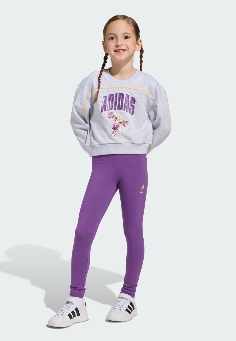 Szara bluza z napisem „ADIDAS” i grafiką postaci, dopasowana do fioletowych legginsów. Białe buty sportowe z czarnymi paskami, gładka faktura, swobodny krój.