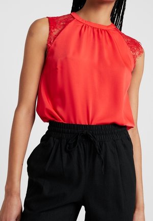 Vrouw draagt een mouwloze rode blouse met kanten schouders en een geraffineerde halslijn, gecombineerd met zwarte broek met trekkoord.